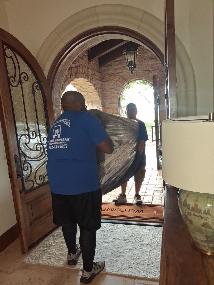 Two Garcia movers carrying a shrink-wrapped piece through an arched front doorway
